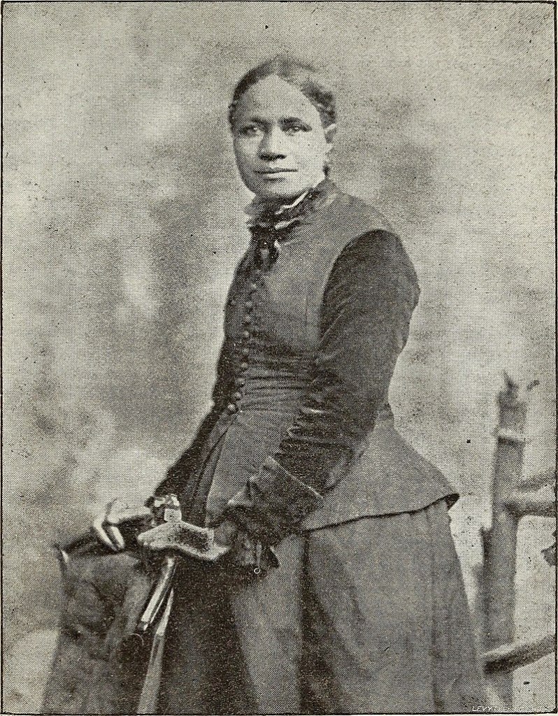 A black and white photo of Frances E. W. Harper. She stands with her body facing to the left, head turned to look at the camera. Her hands rest on a chair in front of her. Her hair is pulled back from her face. She is wearing a dress with a full skirt and long sleeves with wide cuffs. Over the dress she wears a long vest with buttons down the front and a peplum. The high ruffled collar of the dress can be seen above the neckline of the vest.