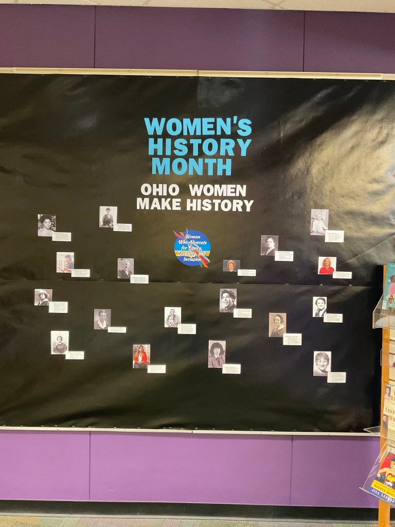 A bulletin board with a black background. Blue words at the top read "Women's History Month" and white words below read "Ohio Women Make History." A colorful logo in the center reads "Women Who Advocate for Equity, Diversity, and Inclusion." There are small photos of women with short bios next to them across the board.